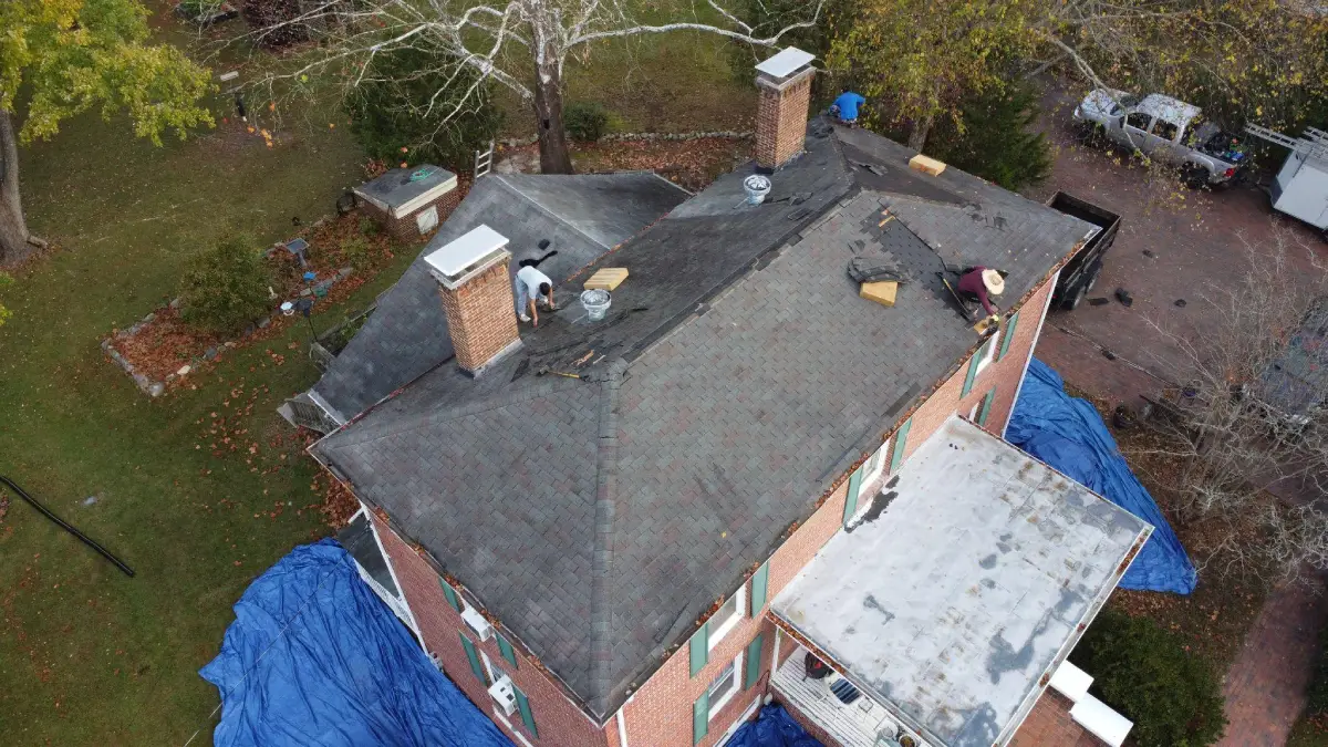 Aerial view of workers on a house roof during repair, with some shingles removed and blue tarps around the building.