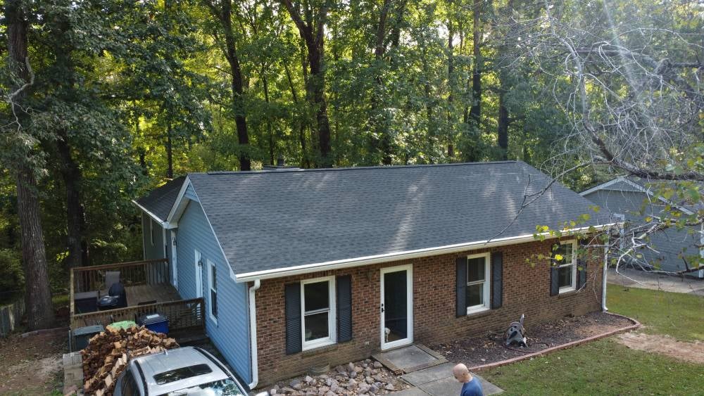 Aerial view of a house with a new black shingle roof, blue siding, brick facade, and a deck with stacked wood.