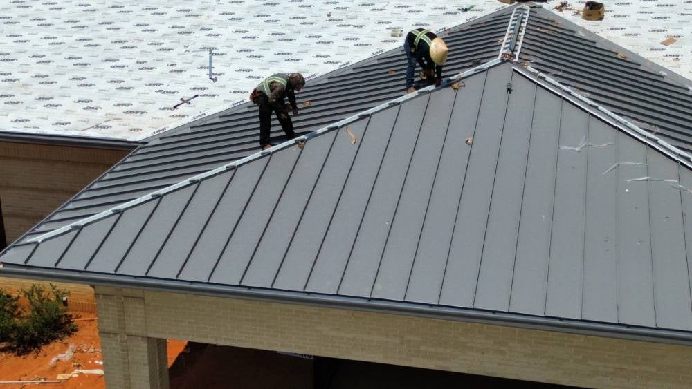 Two workers in safety vests installing dark gray standing seam metal roofing panels on an angled roof.