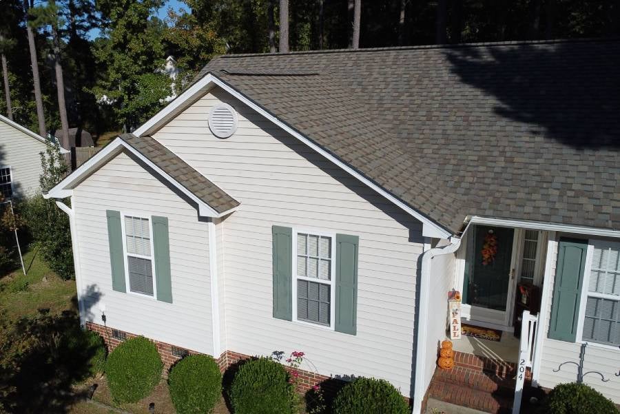 Aerial view of a light-colored house with green shutters, a new gray shingle roof, and a welcoming fall porch display.