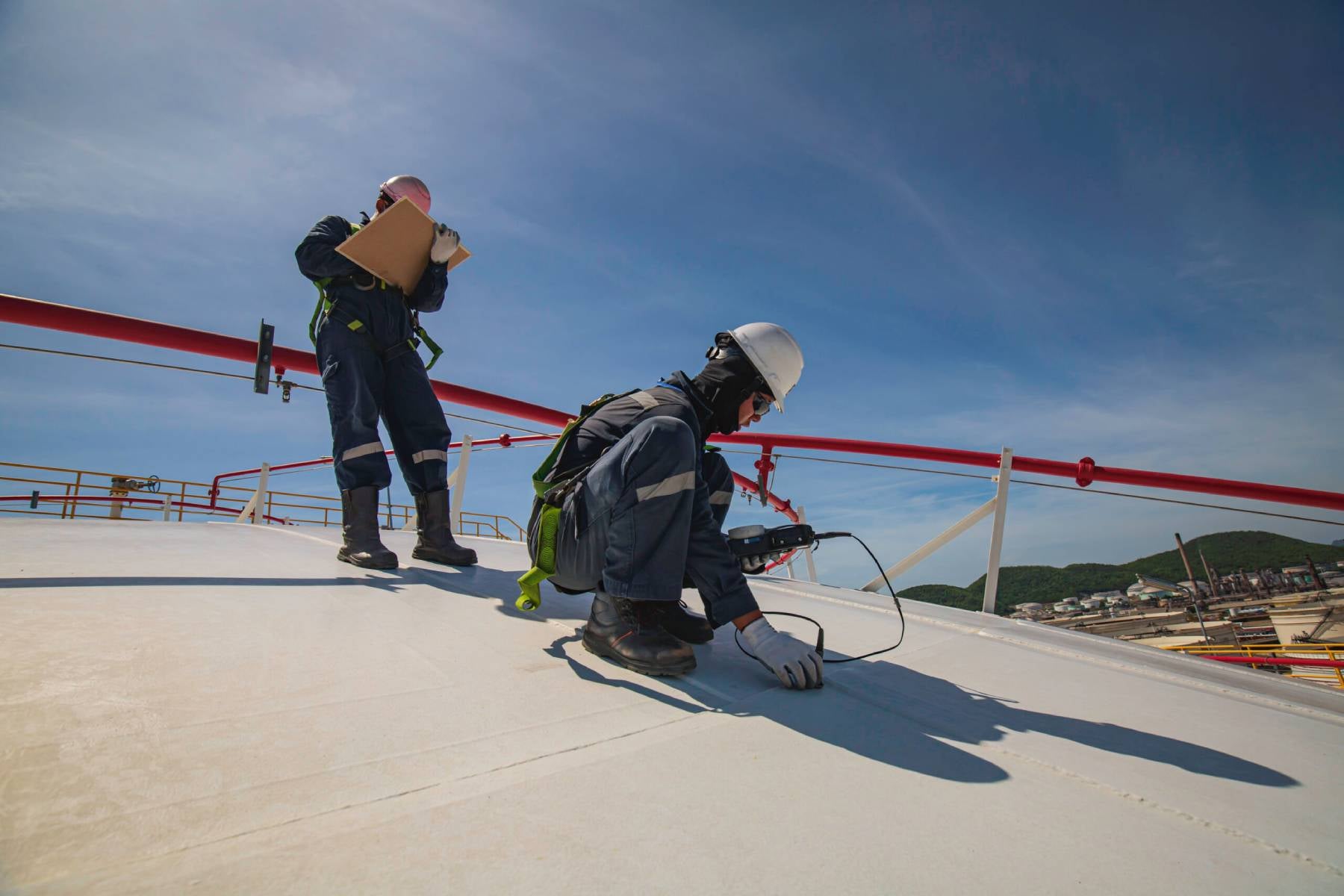 Workers on a large, white tank rooftop, one inspecting with equipment, the other holding a clipboard.