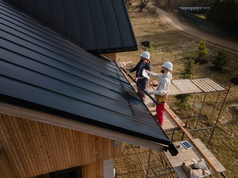 Two workers in hard hats installing dark metal roofing panels on a wooden house, with scaffolding and a dirt road nearby.