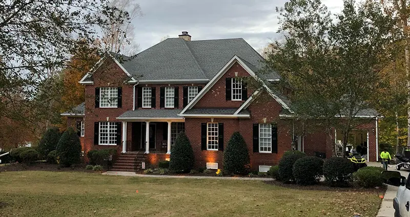 Aerial view of a light-colored house with a textured gray roof, green shutters, autumn decorations, and green bushes.