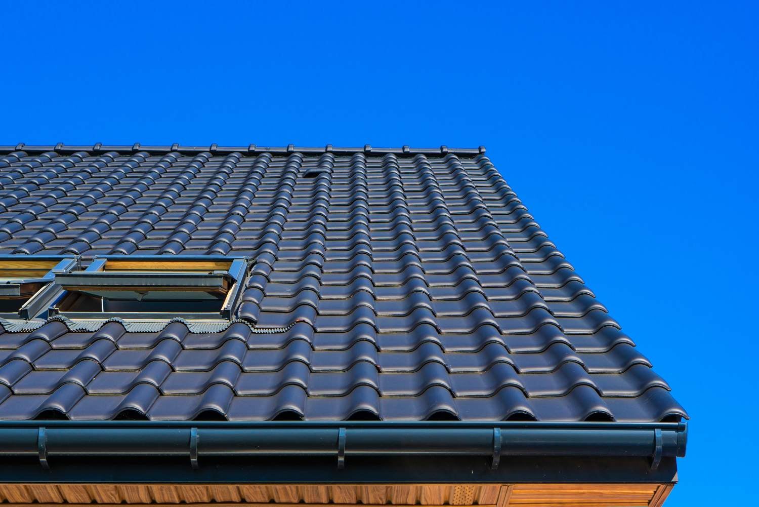 Close-up of a dark tiled roof with two open skylights and a gutter against a clear blue sky.