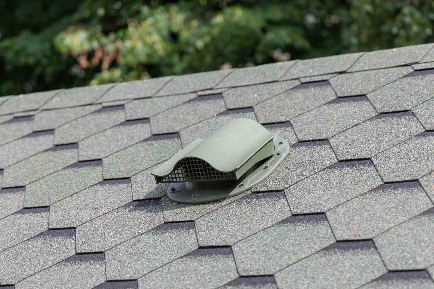 Roofer in a yellow hard hat and safety harness, crouching on a brown shingle roof, repairing it.