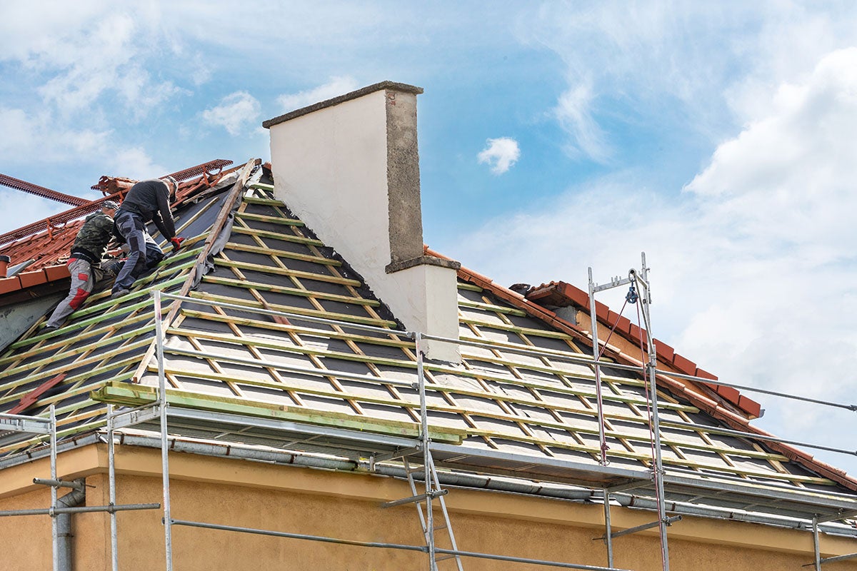 Two roofers work on an unfinished roof under a blue sky, with scaffolding on the side of the house.
