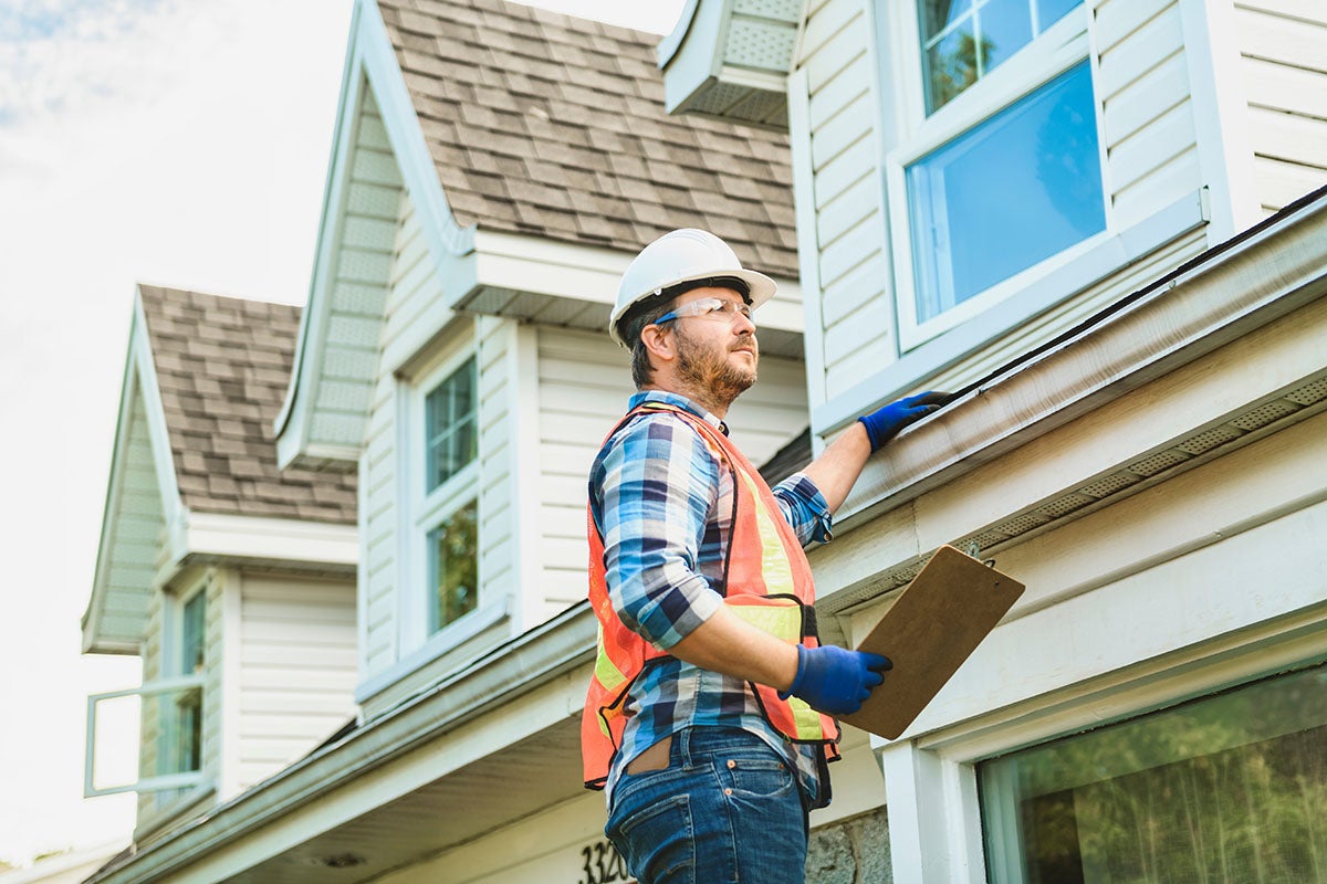 A male home inspector in a hard hat and safety vest checks the gutter on a white house.