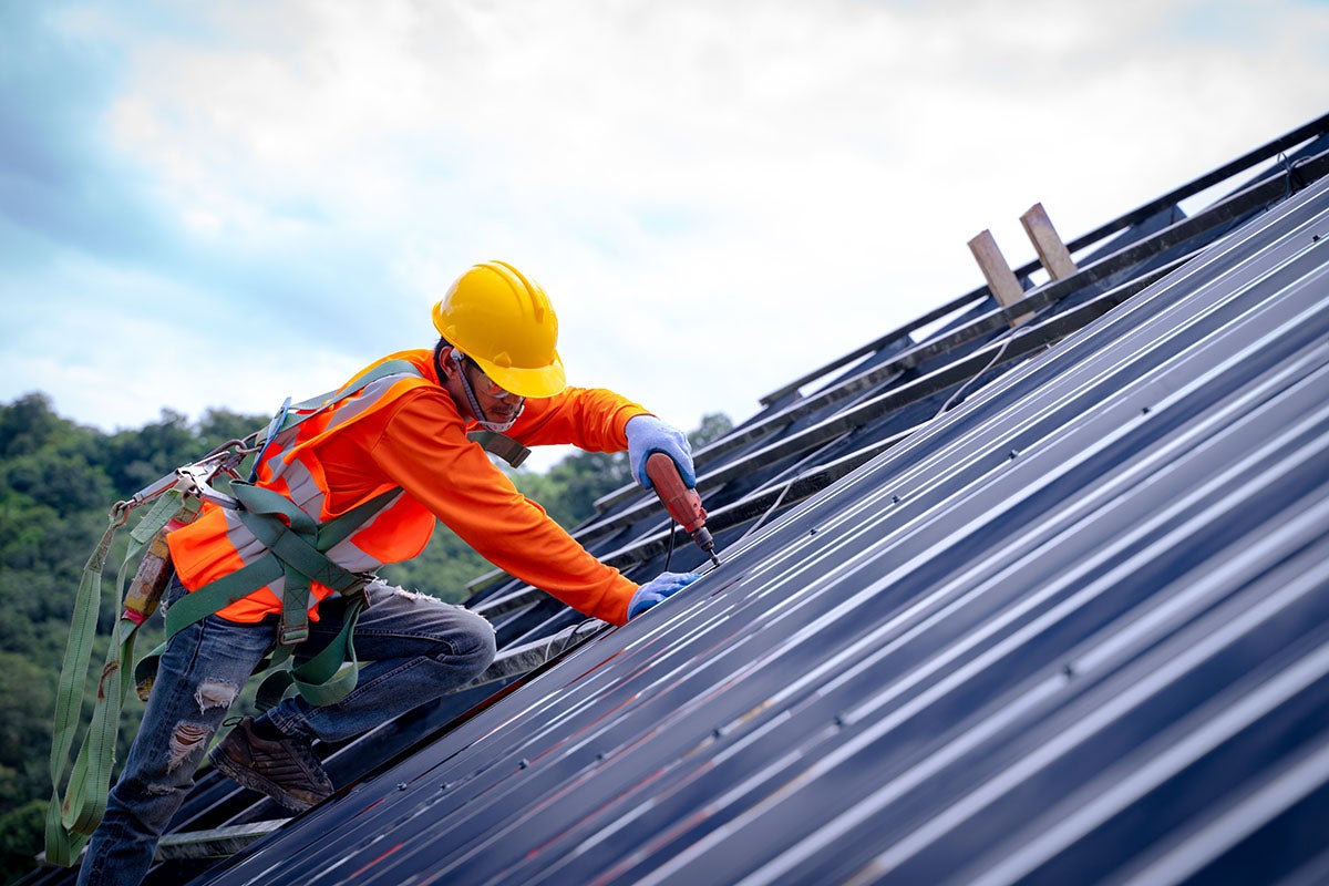 A construction worker in a hard hat and safety harness installs solar panels on a slanted roof.