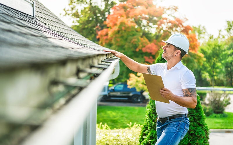 Man in hard hat and white shirt inspecting roof gutter with a clipboard, autumn trees in background.